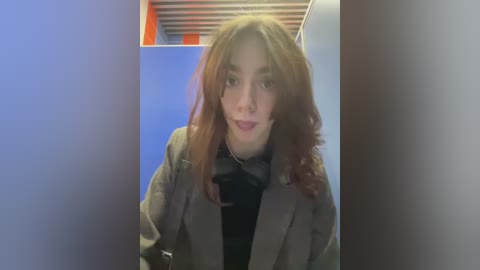 Media: Video of a young woman with shoulder-length auburn hair, wearing a brown blazer over a black turtleneck, standing in a dimly lit hallway with blue and orange walls.