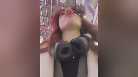 Media: Video of a woman with red hair and fair skin, wearing black headphones, lying on a metal bed in a dimly lit room with pink and blue lights.