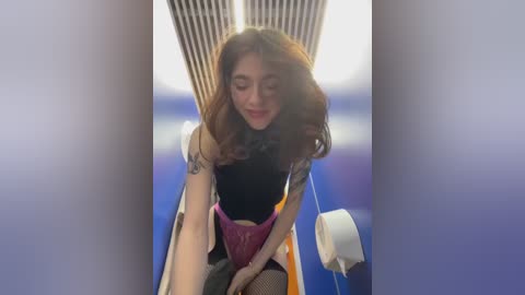 A video of a young woman with long brown hair, wearing a black top, purple lace panties, and fishnet stockings, seated on a blue airplane seat, with a tissue dispenser visible in the background.