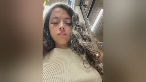 Video of a young woman with long, wavy, dark brown hair, wearing a beige ribbed sweater, sitting in a dimly lit, narrow airplane aisle. She appears contemplative, with a neutral expression.