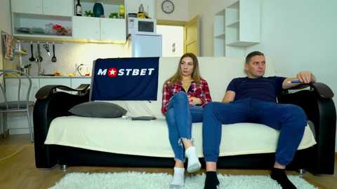 Video of a blonde woman in a plaid shirt and blue jeans sitting on a couch with a man in a navy shirt and jeans, watching TV in a modern, minimalistic living room.