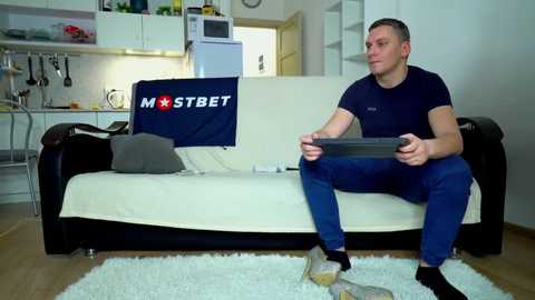 Video of a Caucasian man with short hair, wearing a navy shirt and blue pants, sitting on a white couch with a black cushion, holding a video game controller, in a modern, minimalist living room.