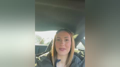 Media: Video of a young woman with light skin and long blonde hair, smiling, seated in a car, wearing a dark jacket, with blurred greenery outside.