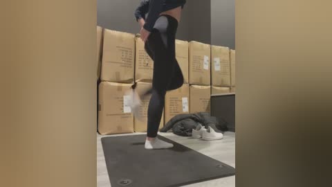 Media: Video of a person in black leggings and a black top, performing a split on a black yoga mat, surrounded by stacked cardboard boxes in a dimly lit room.