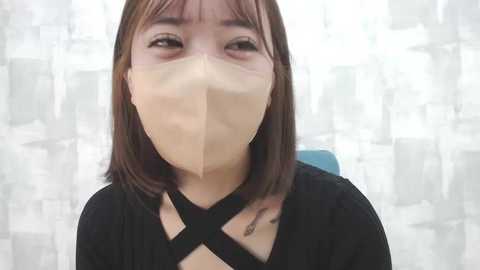 Media: A close-up video of an Asian woman with shoulder-length brown hair, wearing a black shirt with a crisscross design and a beige face mask, set against a blurred, white-and-grey patterned background.