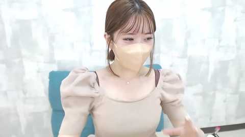 Media: Video of an Asian woman with light skin, straight brown hair, wearing a beige mask, a beige blouse, and dangling gold earrings, seated in front of a blue chair, with semi-transparent white curtains in the background.