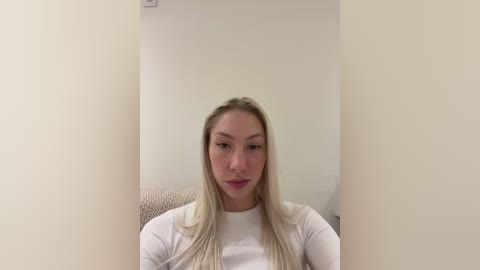 A video of a young, fair-skinned woman with long blonde hair, wearing a white long-sleeve shirt, sitting in a beige chair against a plain white wall, creating a minimalist, neutral background.