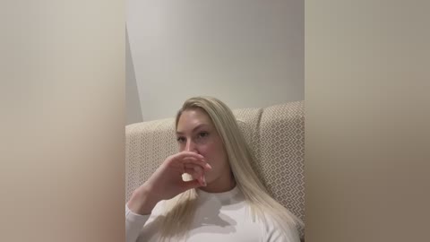 Media: Video of a blonde woman with fair skin, sitting in a beige armchair, wearing a white top. She is blowing her nose, with a tissue in her right hand, against a plain white wall background.