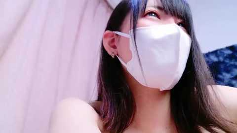 Media: Video of an East Asian woman with long black hair, wearing a white surgical mask, standing against a white and blue background, looking up with a thoughtful expression.