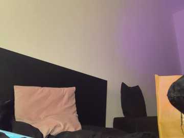 Media: Video of a minimalist bedroom with a black headboard and a single beige pillow on a dark bedspread, against a white wall. A wooden nightstand is partially visible on the right.