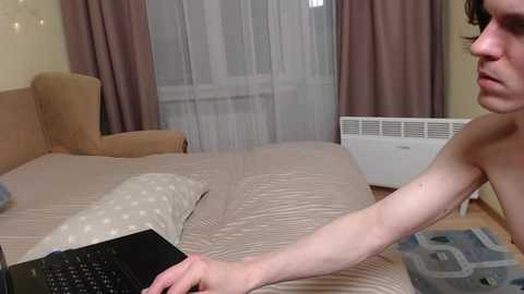 Video of a nude, fair-skinned person with short hair using a laptop on a bed in a room with beige curtains, a brown armchair, and a white radiator.