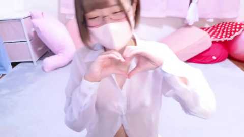 Media: Video of an Asian woman with glasses, wearing a white surgical mask, making heart shapes with her hands in front of a pink bed with plush pillows.