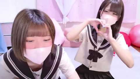 Media: Video of two young Asian girls wearing matching school uniforms with white masks, making heart shapes with their hands in a brightly lit room with pastel decor.