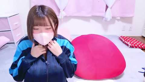 Media: Video of a young Asian woman with straight brown hair and light skin, wearing a blue and black jacket, a white face mask, and a red heart-shaped pillow on a light-colored floor, in a pink-themed room.