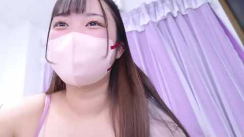 Media: Video of an East Asian woman with long brown hair, wearing a light pink face mask and spaghetti straps, standing in a softly lit room with lavender curtains.