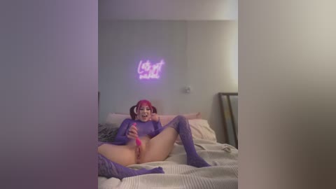 Video of a young, fair-skinned woman with pink hair, wearing a purple bodysuit and knee-high socks, lying on a bed in a dimly-lit room, masturbating with a pink vibrator.