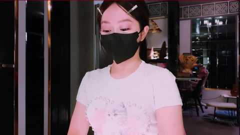 Media: Video of an East Asian woman wearing a black face mask, white top, and hair clips, standing indoors in a modern, dimly lit room with a glass window, a desk, and a chair.