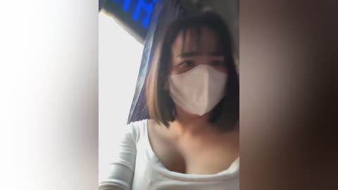 Media: Video of an Asian woman with medium-length black hair, wearing a white face mask and a white top, standing in a dimly lit room with blurred blue and brown walls.