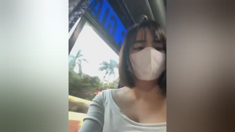 Media: Video of a young woman with short dark hair, wearing a white face mask, gray top, standing in a bus with a blue banner, palm trees, and greenery outside the window.