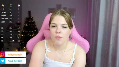 Media: A video of a young woman with fair skin and light brown hair, wearing a white spaghetti-strap top, sitting in a pink gaming chair. She looks serene with closed eyes. Background shows a decorated Christmas tree and soft lighting.