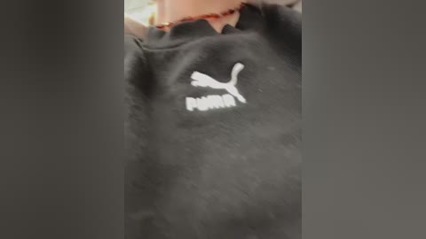 Video of a black Puma sweatshirt with a white logo and \"Puma\" text, partially obscured by a person's arm, possibly a child. Background is blurry and out of focus.