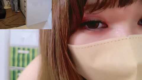 Media: Video of a person's face with brown hair partially covering the eye, wearing a beige surgical mask, with blurred background featuring a green and white striped storage box.