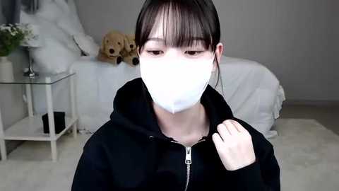 Media: A video of a young Asian woman with long black hair, wearing a white surgical mask and black hoodie, standing indoors near plush teddy bears and a glass table with a vase.