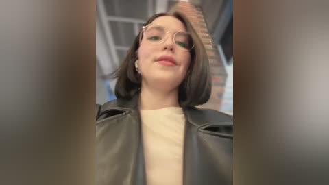 Media: Video of a young woman with shoulder-length brown hair, wearing glasses and a black leather jacket over a cream-colored top, standing in an indoor setting with blurred architectural elements in the background.