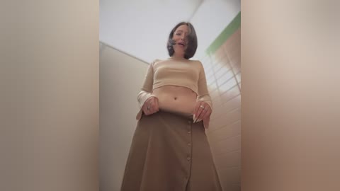 Media: A video of a slender, light-skinned woman with short brown hair, wearing a beige crop top and high-waisted skirt, standing in a narrow, beige hallway with a blurred green object in the background.