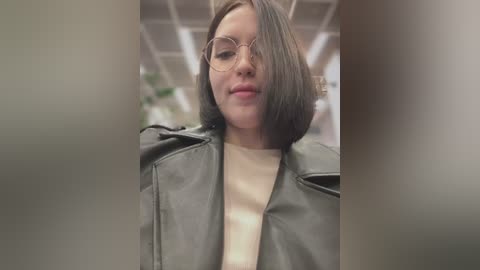 Media: A video of a young woman with short, straight black hair, wearing glasses, a beige sweater, and a black leather jacket, taken in a modern indoor setting with fluorescent lighting.