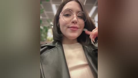 Media: Video of a young woman with light skin, glasses, and brown hair, wearing a black leather jacket, in a green-lit room.