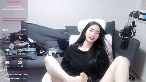 Media: Video of an Asian woman with long black hair, wearing a black sweater, sitting on a grey couch, holding a microphone, in a minimalist living room with grey walls and a black lamp.