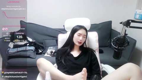 Media: A video of an East Asian woman with long black hair, wearing a black top, reclining on a white chair, surrounded by a black couch, clothes, and a microphone.