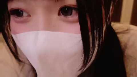 Media: Video of a young Asian woman with long black hair, wearing a white surgical mask, covering her mouth and nose. Her large, dark eyes are visible, with a neutral expression. The background is blurred, suggesting an indoor setting.