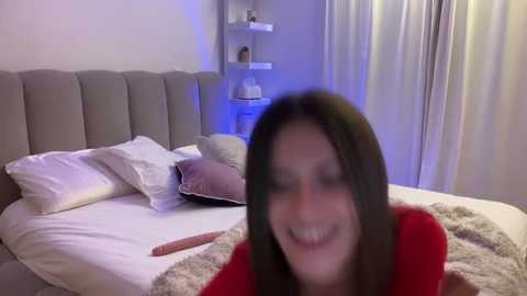 Media: Video of a woman with long brown hair, smiling, lying on a bed with white linens, beige pillows, and a plush blanket. Background features a modern, minimalist bedroom with a light-colored upholstered headboard and white curtains.