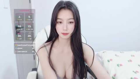 Media: A video of an East Asian woman with long black hair, fair skin, and red lipstick, wearing a revealing black top, sitting on a bed with a floral duvet. A digital health tracker display shows daily stats on the left.