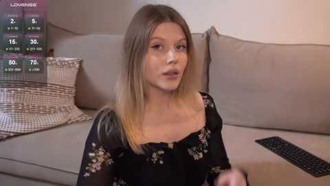 Video of a young Caucasian woman with light skin and straight blonde hair, wearing a black floral dress, seated on a beige couch in a living room.