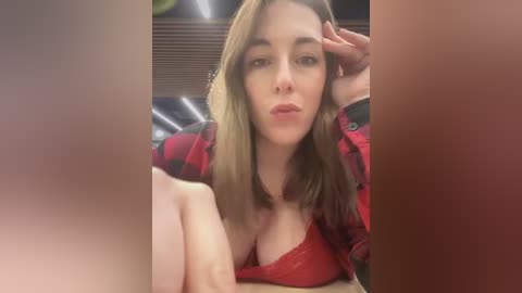 Media: A video of a young Caucasian woman with straight, light brown hair, wearing a red lace bra and a plaid jacket. She has a relaxed, casual pose, with a hand on her head. The background is blurred, featuring muted tones and a wooden surface.