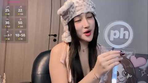 Media: Video of a smiling Asian woman with long black hair, wearing a fuzzy white hat, pink top, and holding a phone with a \"ph\" logo.