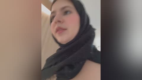 A close-up video of a young woman with fair skin, wearing a black hijab and a slight blush on her cheeks, gazing upwards. The background is blurred, featuring beige and dark brown tones.