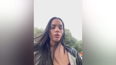 Media: Video of a young woman with long, straight dark hair, wearing a black leather jacket, standing outdoors with a blurred green background.