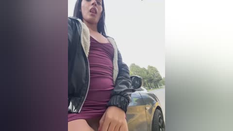 Media: Video of a Latina woman with long black hair, wearing a tight maroon dress and black leather jacket, sitting in a car with a blurred background of trees and a yellow vehicle.