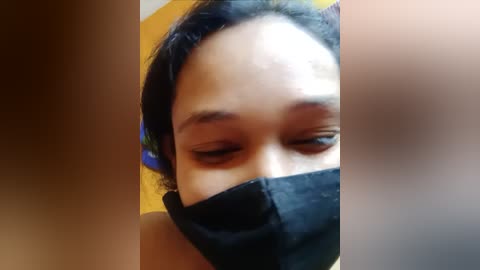 Media: Video of a woman with medium-brown skin and short, dark hair, wearing a black face mask. Her eyes are closed, and she is partially visible from the neck up. The background is blurry, with warm colors.