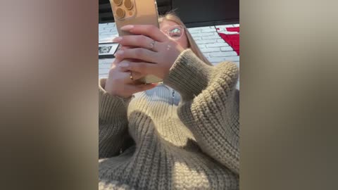 Media: A video of a woman with glasses taking a selfie indoors, wearing a beige knit sweater, against a brick wall background.