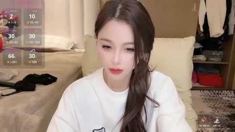 Media: Video of a young Asian woman with long dark hair, fair skin, and red lipstick, wearing a white T-shirt, sitting on a beige sofa. Background includes a wooden shelf, clothes, and a virtual overlay showing health stats.