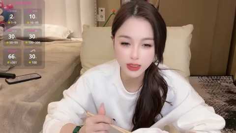 Media: A video of an East Asian woman with fair skin, long black hair, and a white sweatshirt, sitting on a beige couch, holding a phone. The background shows a cluttered living room with a TV and a bed.