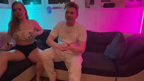 Media: Video of two young adults, a blonde woman and a man, sitting on a dark blue couch. The room is dimly lit with neon pink and blue lights, creating a modern, lively atmosphere.