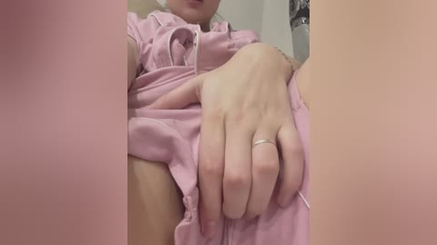 Video of a light-skinned person in pink scrubs, with a visible hand on their genital area, wearing a silver ring, in a dimly lit room.