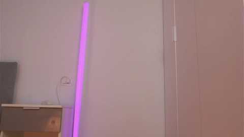 A video of a minimalist room with a vertical purple LED strip light on a white wall, a flat-screen TV on a wooden stand, and a white door to the right.