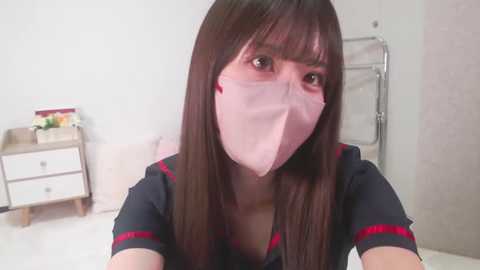 Media: A video of an Asian woman with long, straight black hair, wearing a black nurse uniform with red trim and a pink surgical mask. She stands in a minimalist room with white walls, a metal rack, and a bedside table.